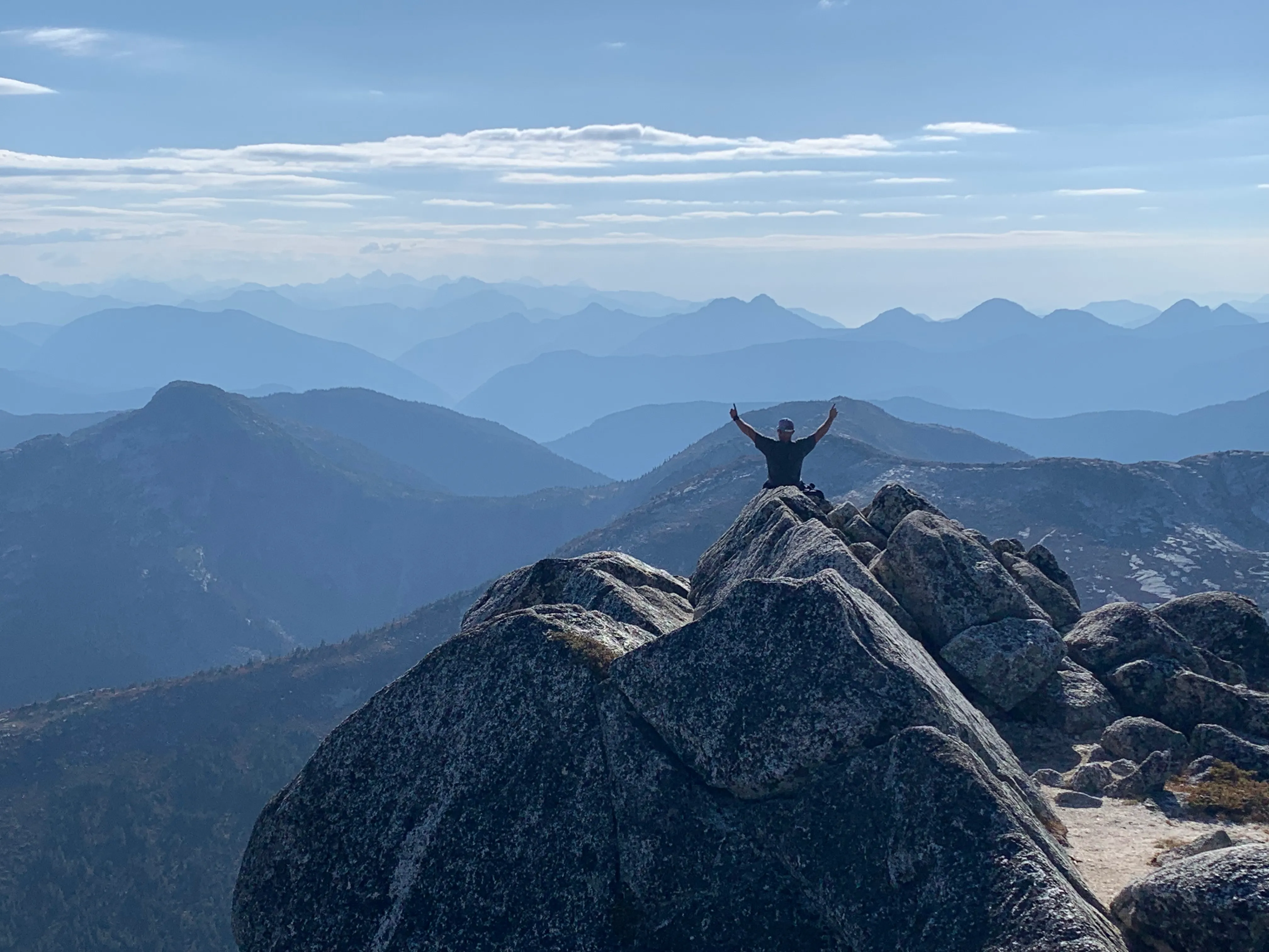 Trevor Rizun on a rocky summit, arms raised, blue ridgelines behind